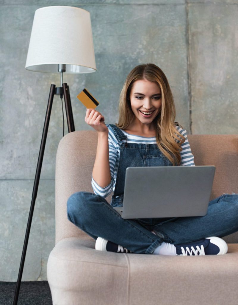 happy beautiful woman holding credit card in hand and sitting on sofa with laptop